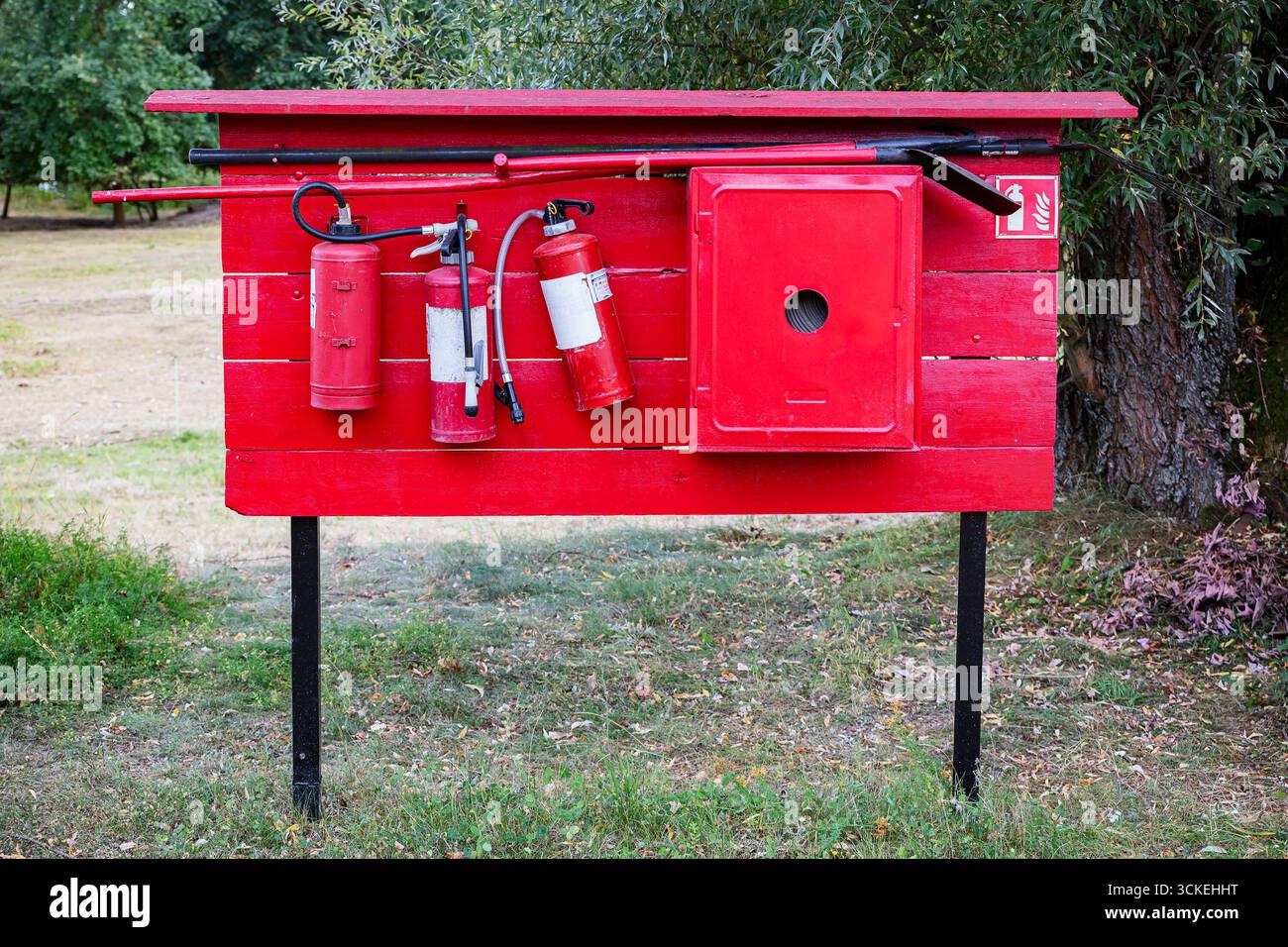Poste de sécurité incendie extérieur rouge vif avec plusieurs extincteurs, un tuyau, une pelle, un crochet et une armoire métallique montés sur une planche de bois dans un parc Banque D'Images