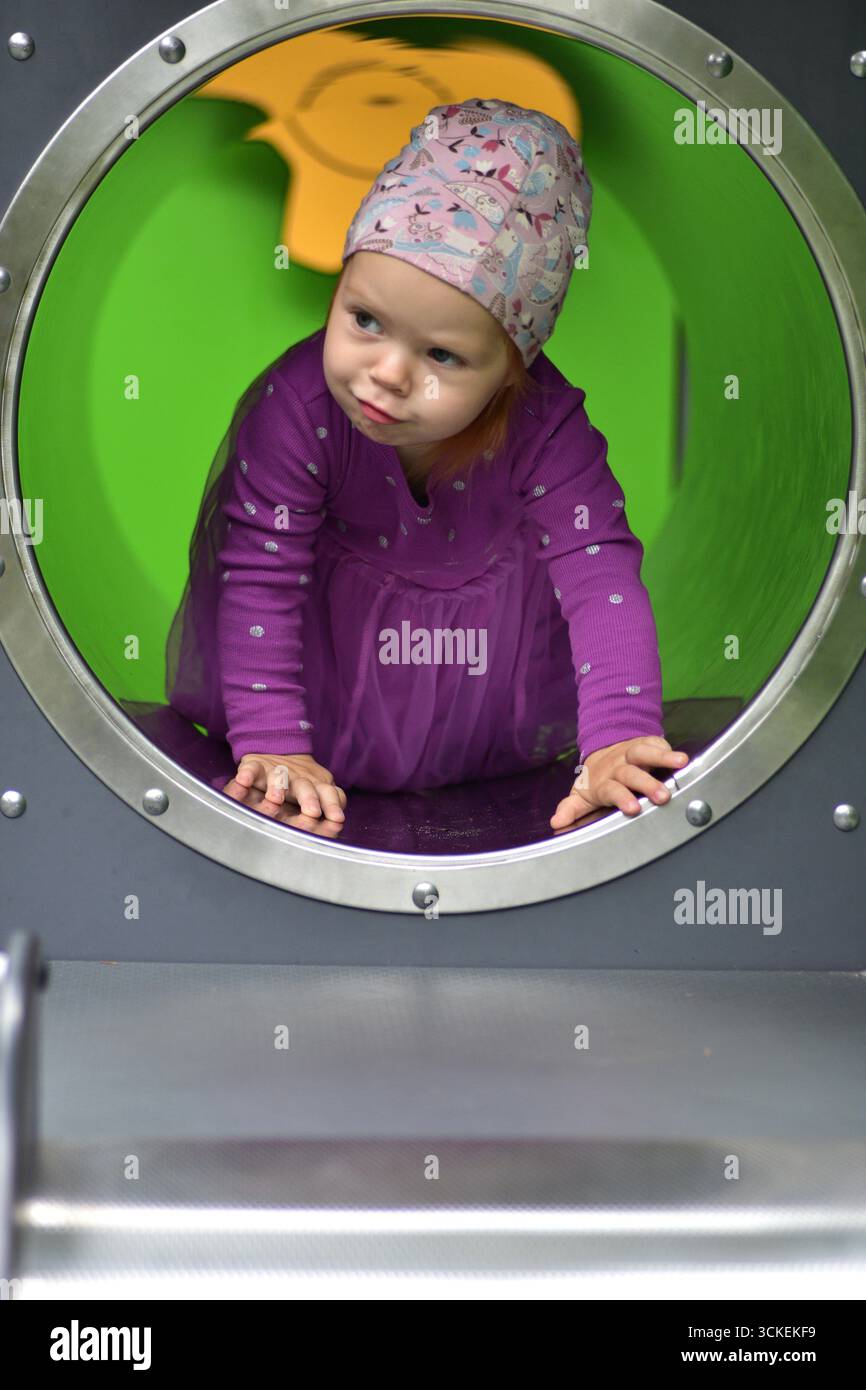 Mignon jeune fille en robe violette et chapeau à motifs rampant à l'intérieur du tunnel vert de terrain de jeu. Enfant explorant la structure de jeu avec curiosité et plaisir exp Banque D'Images