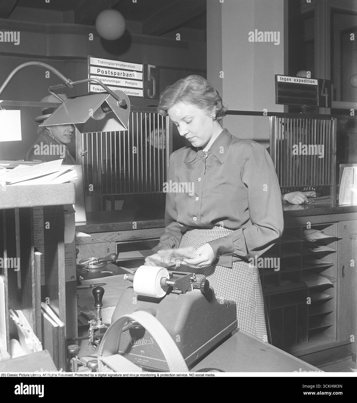 Intérieur d'une banque avec une jeune femme derrière le comptoir dans le bureau de la banque. Elle est vue en train d'aider un client qui est vu debout au comptoir des caisses. L'image symbolise l'histoire financière et la culture de l'épargne personnelle, évoquant la nostalgie des pratiques bancaires traditionnelles. 1950. Kristoffersson ref AY6-3 Banque D'Images