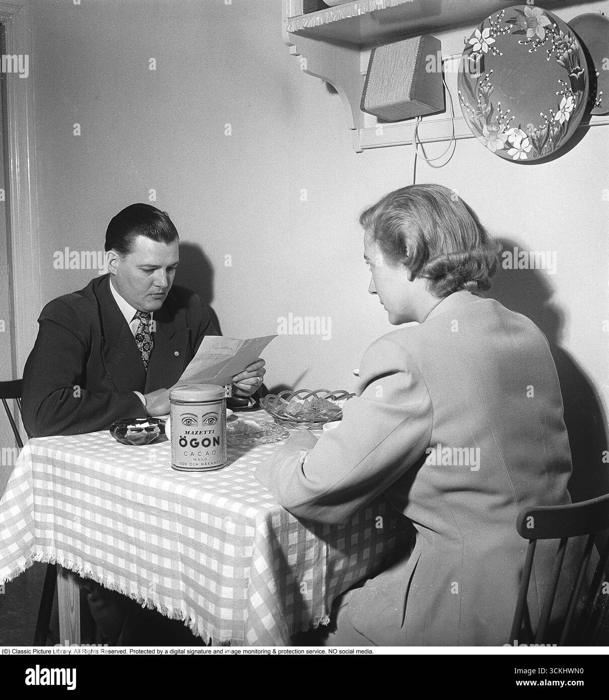 Couple à une table dans les années 1950 Un couple est assis en face l'un de l'autre à une petite table à manger, buvant du chocolat chaud à base de cacao Mazetti Ögon. La boîte, avec son design emblématique des yeux, est un morceau de l’histoire de l’emballage et un élément clé du succès de Mazetti. « Voyez de vos propres yeux que vous obtenez les yeux de Mazetti » était un slogan publicitaire populaire. Mazetti, une marque de confiserie suédoise de premier plan, est devenue synonyme de cacao de qualité, et sa boîte de cacao Ögon, avec son motif d'yeux distinctif, reste un symbole nostalgique du design suédois du milieu du XXe siècle. 1951. Kristoffersson ref BC51-10. Banque D'Images
