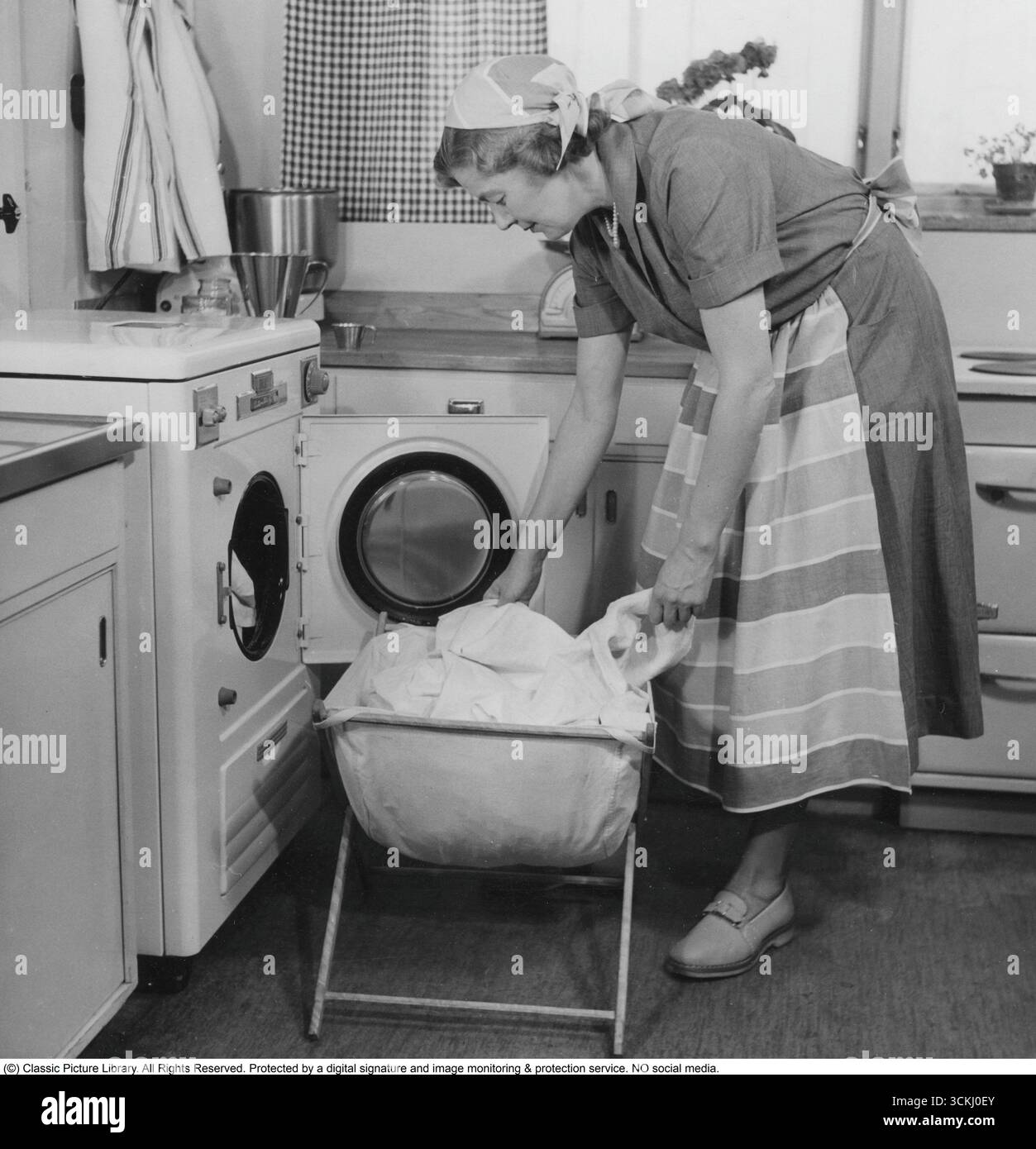 Faire la lessive dans les années 1950 Une femme dans la cuisine met des vêtements sales dans la machine à laver. Elle porte un tablier. 1955 Banque D'Images