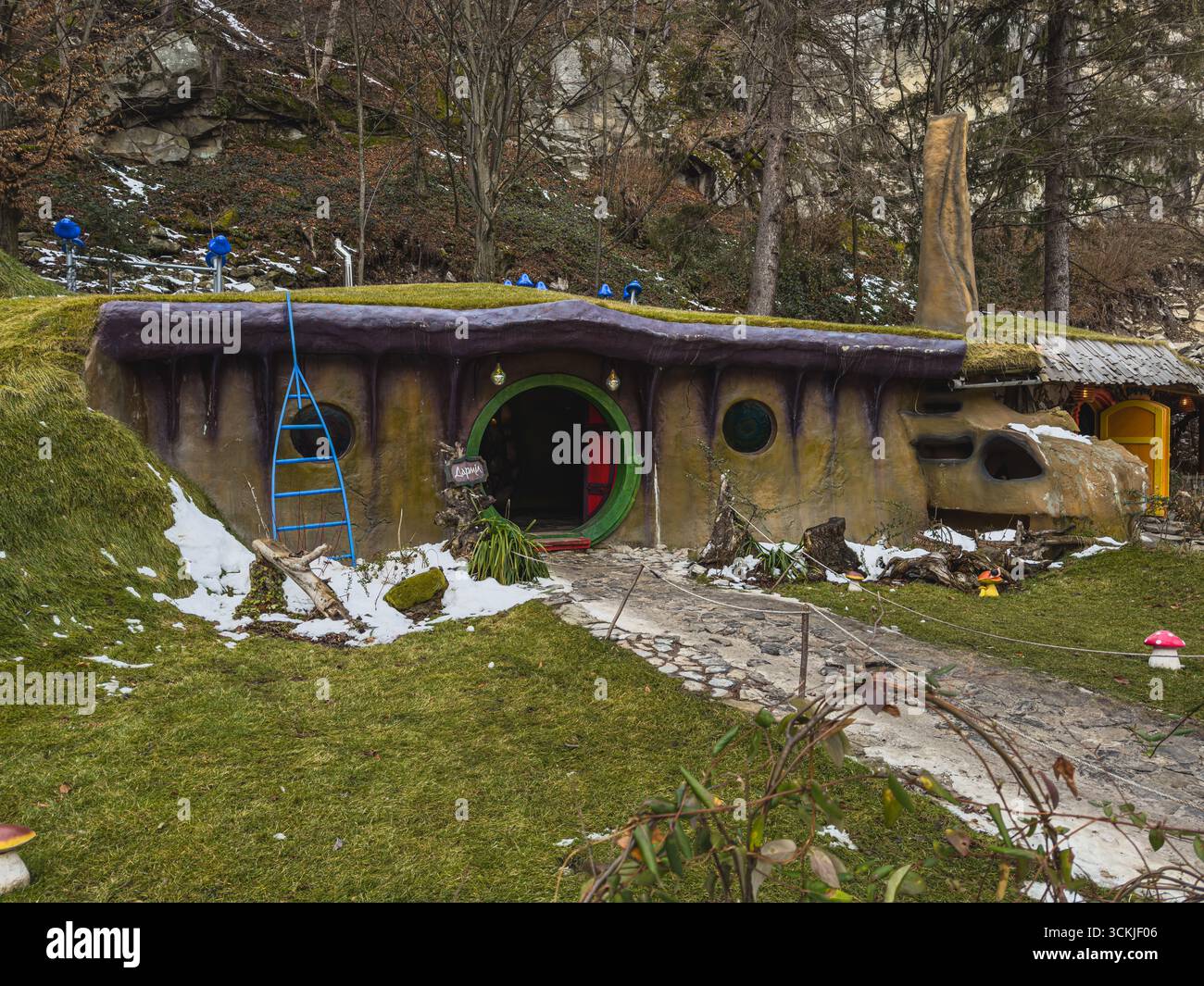 Une maison fantaisiste avec des caractéristiques vibrantes se trouve au milieu d'une forêt enneigée. L'herbe verte et la neige éparpillée créent un cadre hivernal serein, complété par le tre Banque D'Images