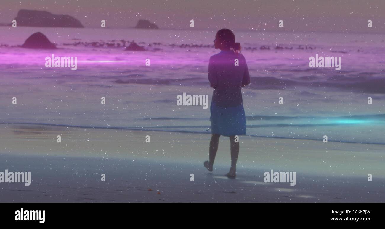 Femme debout pieds nus en robe bleue face à la mer sur la plage au crépuscule, bande violet-bleu et rochers Banque D'Images