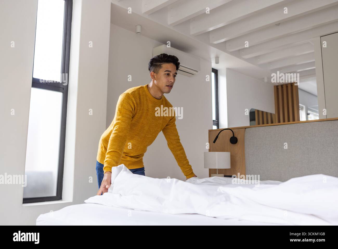 Homme asiatique faisant lit dans une chambre lumineuse avec draps blancs, tête de lit grise, unité split AC Banque D'Images