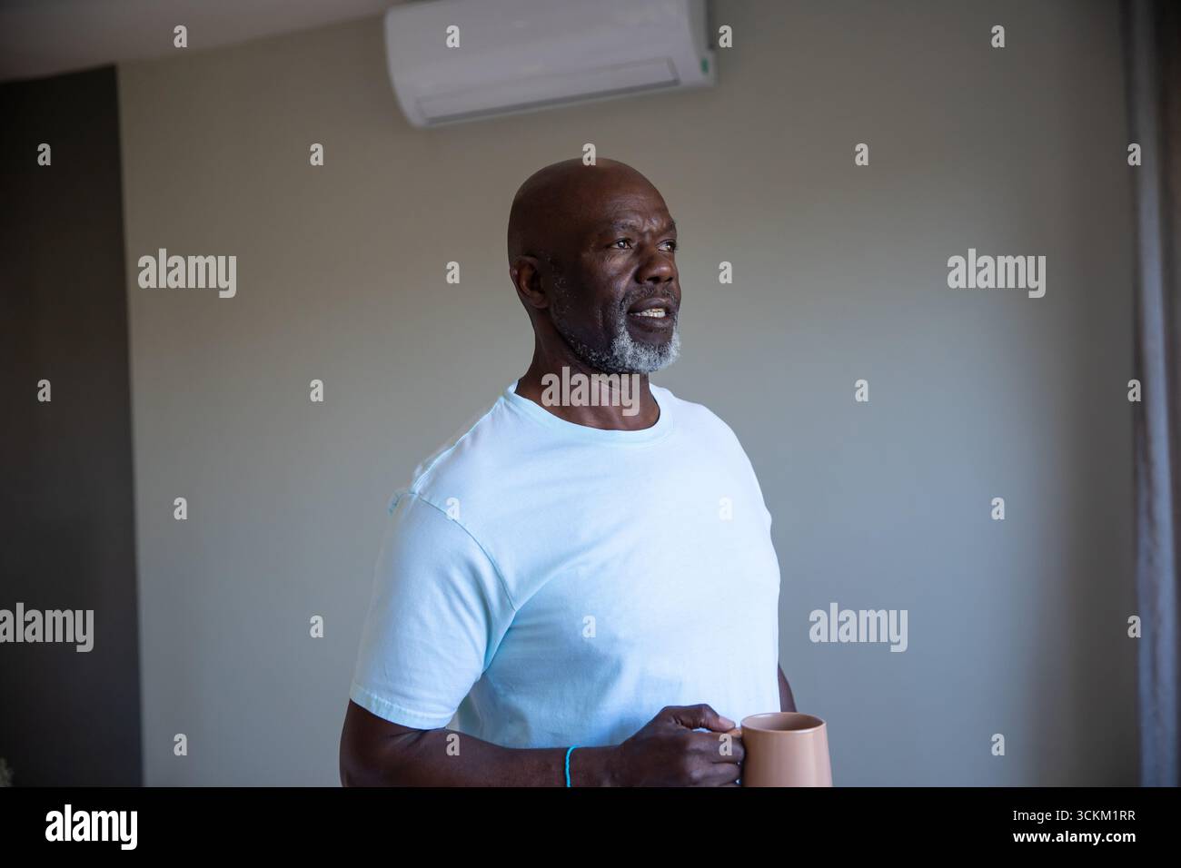Homme afro-américain senior debout à la maison tenant une tasse beige, regardant l'unité AC, espace de copie Banque D'Images