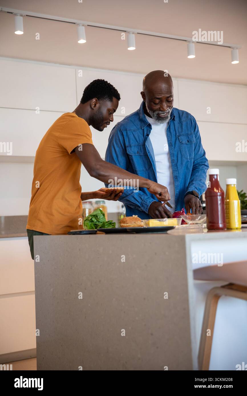 Père afro-américain et fils préparant des hamburgers à l'îlot de cuisine avec couteau, brioches, condiments Banque D'Images