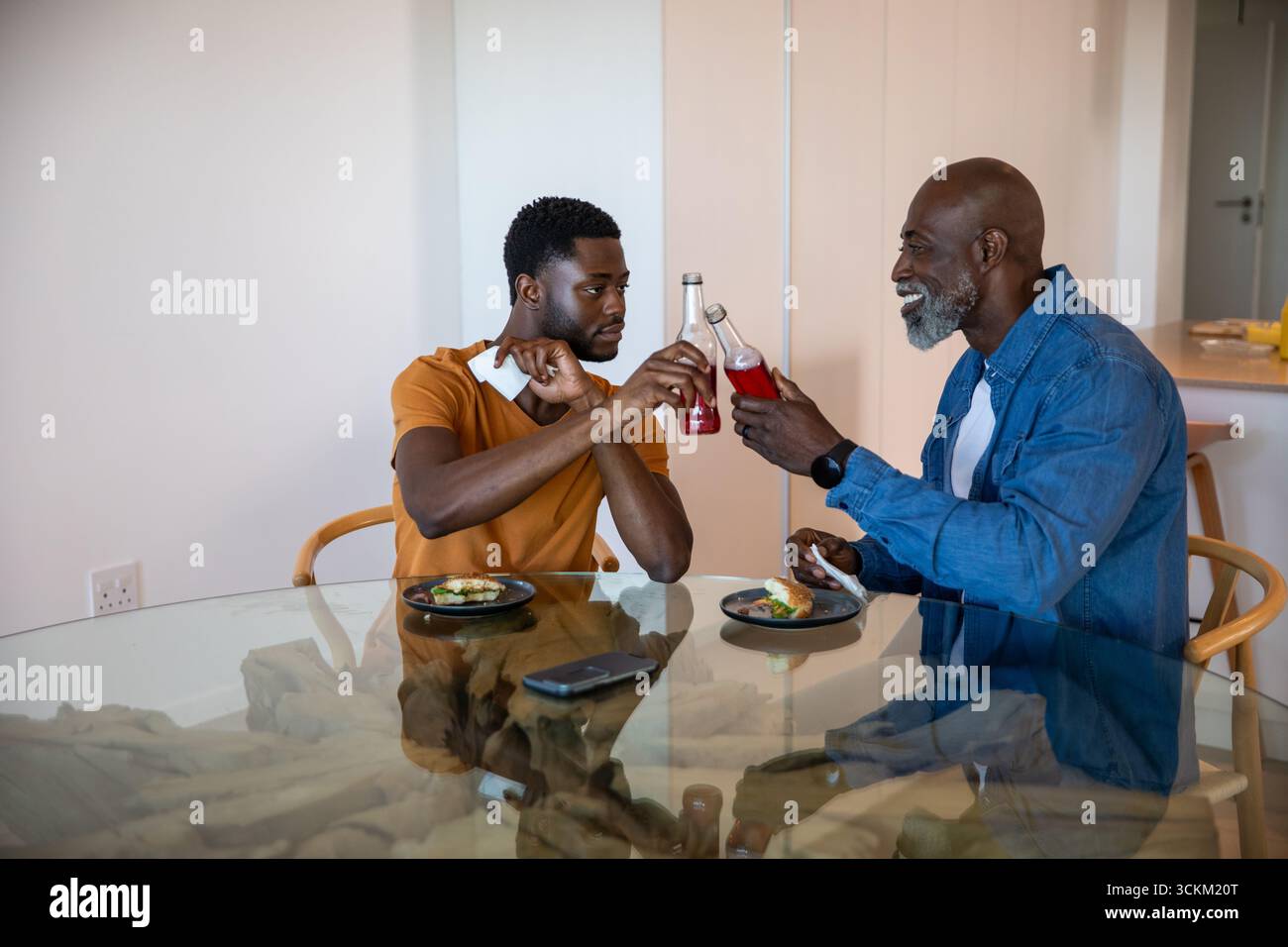 Père et fils afro-américains serrant des bouteilles de boisson rouges à table avec smartphone Banque D'Images