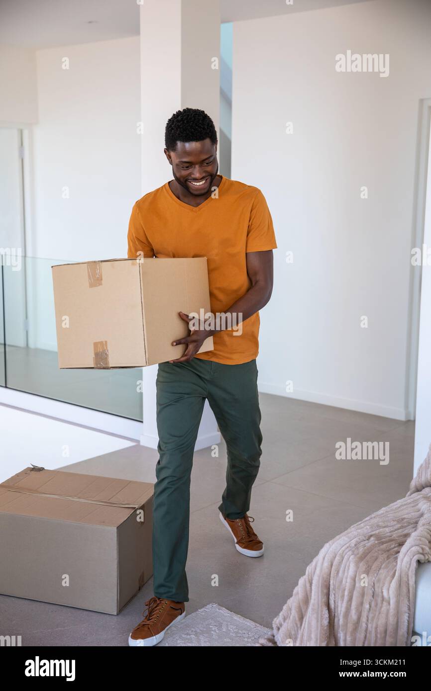 Homme afro-américain transportant une boîte en carton collée à travers l'espace de vie avec garde-corps en verre Banque D'Images Homme afro-américain transportant une boîte en carton collée à travers l'espace de vie avec garde-corps en verre Banque D'Images