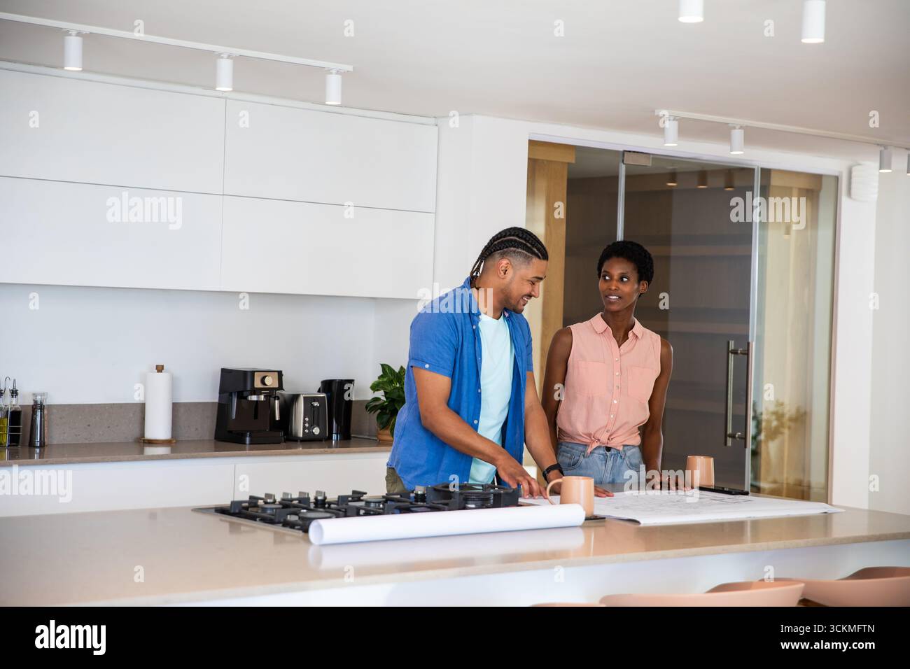 Couple diversifié examinant les plans sur l'îlot de cuisine près des tasses à café et de la table de cuisson à gaz Banque D'Images