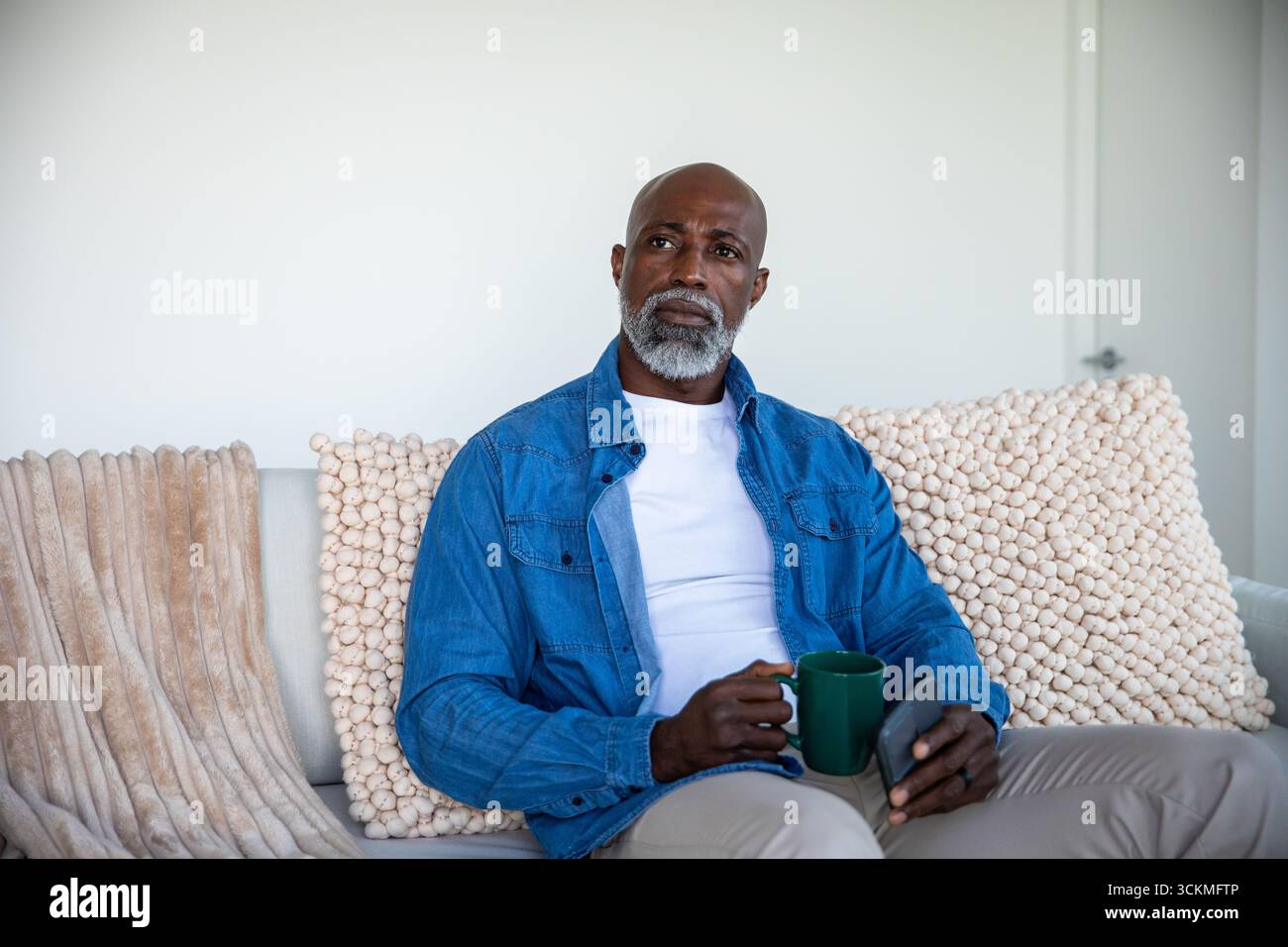 Homme afro-américain senior assis sur le canapé dans le salon tenant la tasse verte, le téléphone défilant Banque D'Images Homme afro-américain senior assis sur le canapé dans le salon tenant la tasse verte, le téléphone défilant Banque D'Images