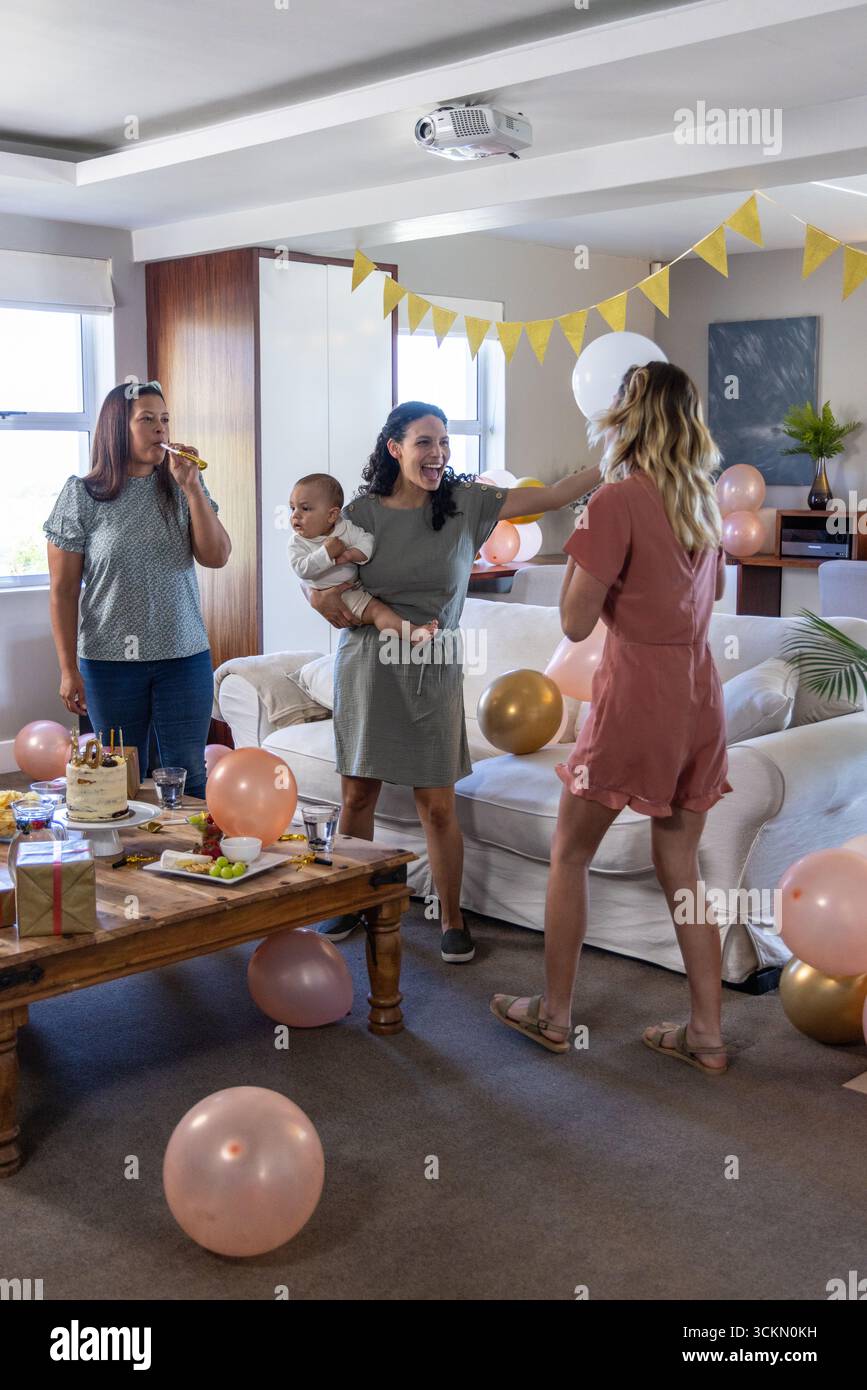 Diverses amies féminines se réunissant dans le salon autour de la table avec gâteau, cadeau enveloppé et ballons Banque D'Images