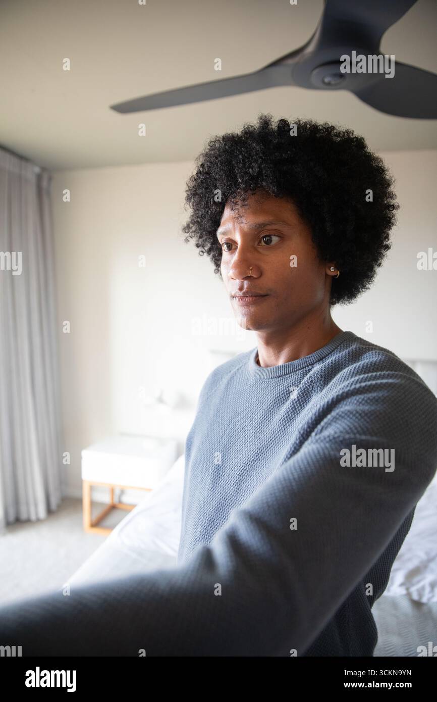 Homme afro-américain debout à côté du lit dans un pull gris avec table de chevet, lampe dans la chambre Banque D'Images Homme afro-américain debout à côté du lit dans un pull gris avec table de chevet, lampe dans la chambre Banque D'Images
