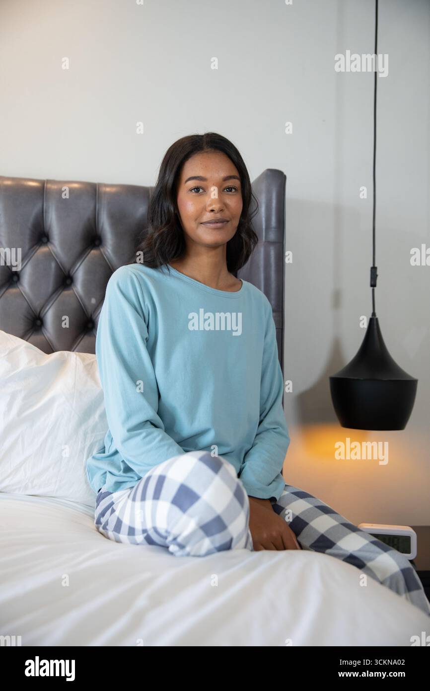 Femme afro-américaine assise dans la chambre sur le lit touffeté en pyjama bleu à côté de la lampe et de l'horloge Banque D'Images Femme afro-américaine assise dans la chambre sur le lit touffeté en pyjama bleu à côté de la lampe et de l'horloge Banque D'Images