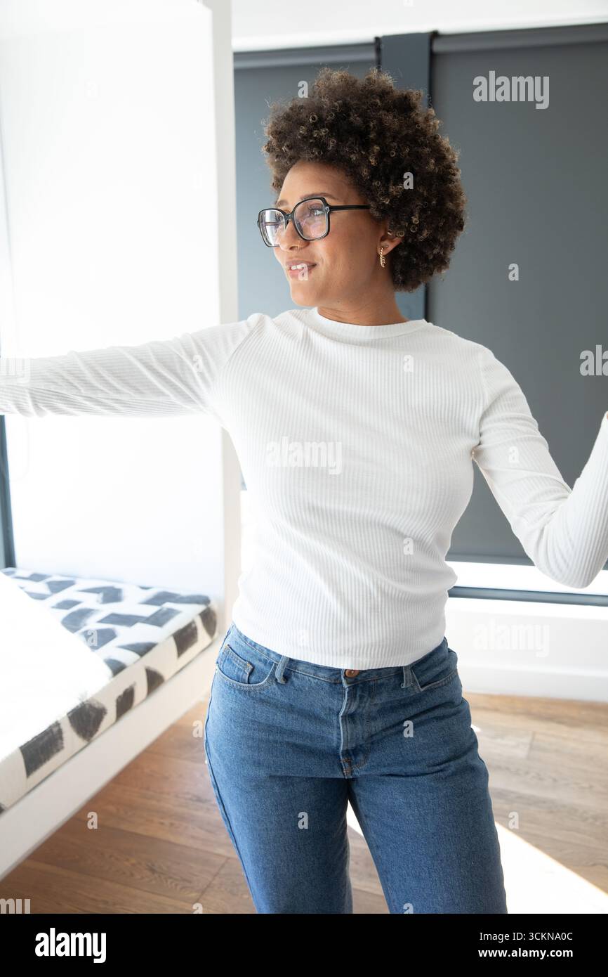 Femme afro-américaine debout dans la chambre atteignant la fenêtre près du lit et l'abat-jour Banque D'Images