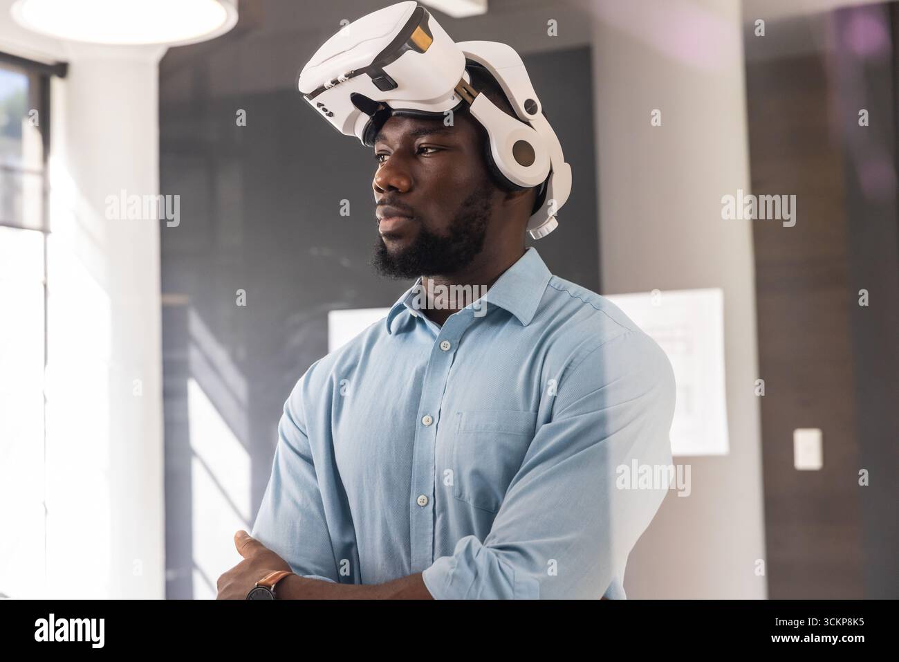Homme afro-américain debout dans le bureau par fenêtre avec la lumière du jour, portant casque vr, montre-bracelet Banque D'Images