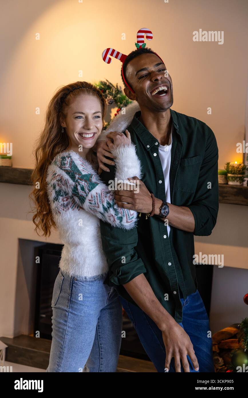 Couple diversifié embrassant, riant dans le salon décoré par cheminée mantel avec des bougies allumées Banque D'Images
