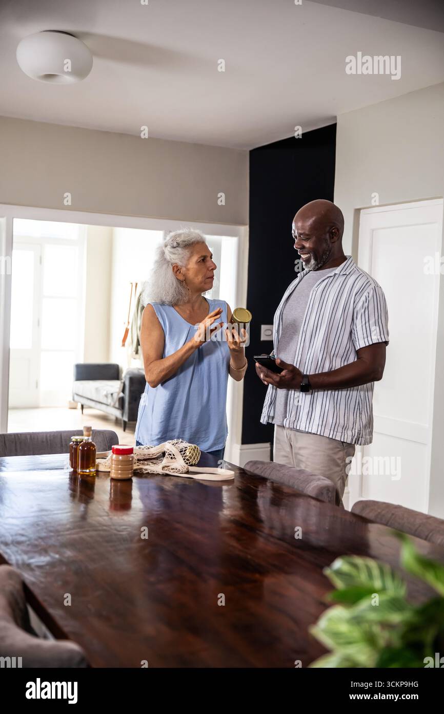 Couple diversifié debout à la table à manger tenant un sac en filet et des pots à condiments tout en utilisant un smartphone Banque D'Images