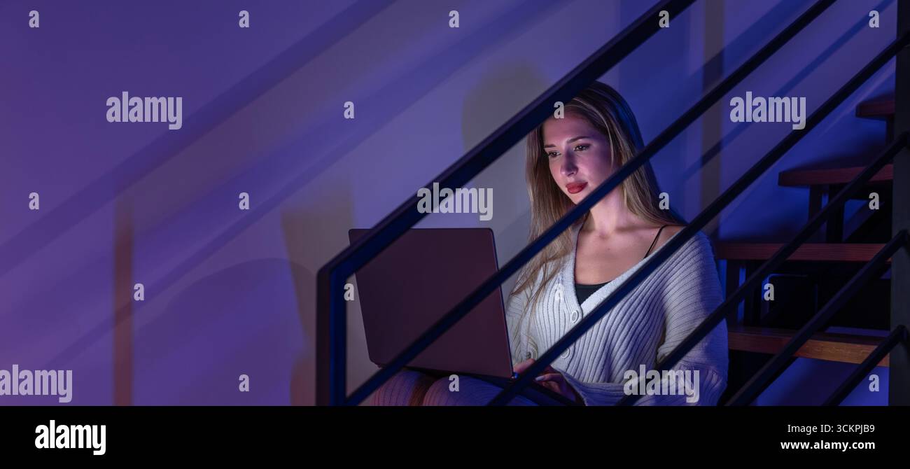 Jeune femme avec ordinateur portable en néon bleu travaillant dans les escaliers. Bureau à domicile de nuit. Banque D'Images