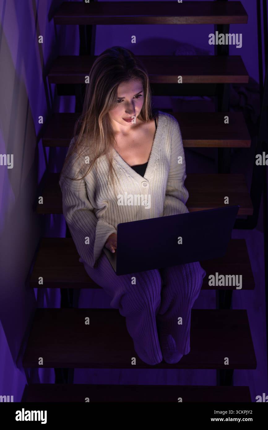 Jeune femme dans les escaliers avec ordinateur portable sous néon la nuit. Ambiance de bureau à domicile. Banque D'Images