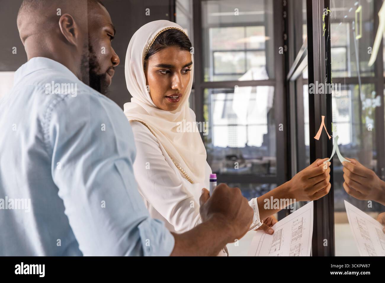 Divers collègues collaborant sur les plans d'étage à la cloison en verre du bureau avec des notes adhésives Banque D'Images