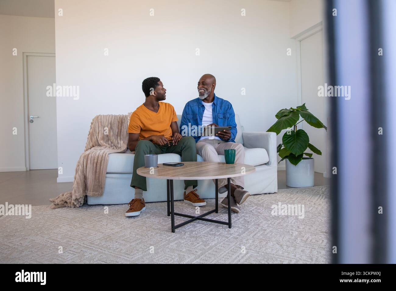 Père afro-américain et fils discutant sur tablette, tasses à café dans le salon Banque D'Images Père afro-américain et fils discutant sur tablette, tasses à café dans le salon Banque D'Images