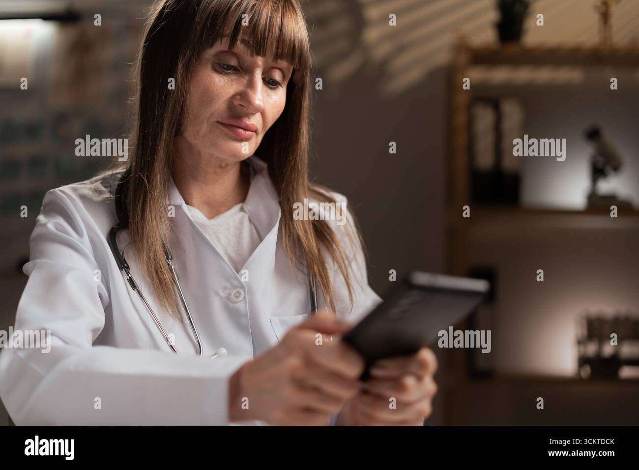 Femme d'âge moyen médecin en manteau blanc assis à table et à l'aide d'un smartphone pendant le quart de nuit. Copier l'espace Banque D'Images