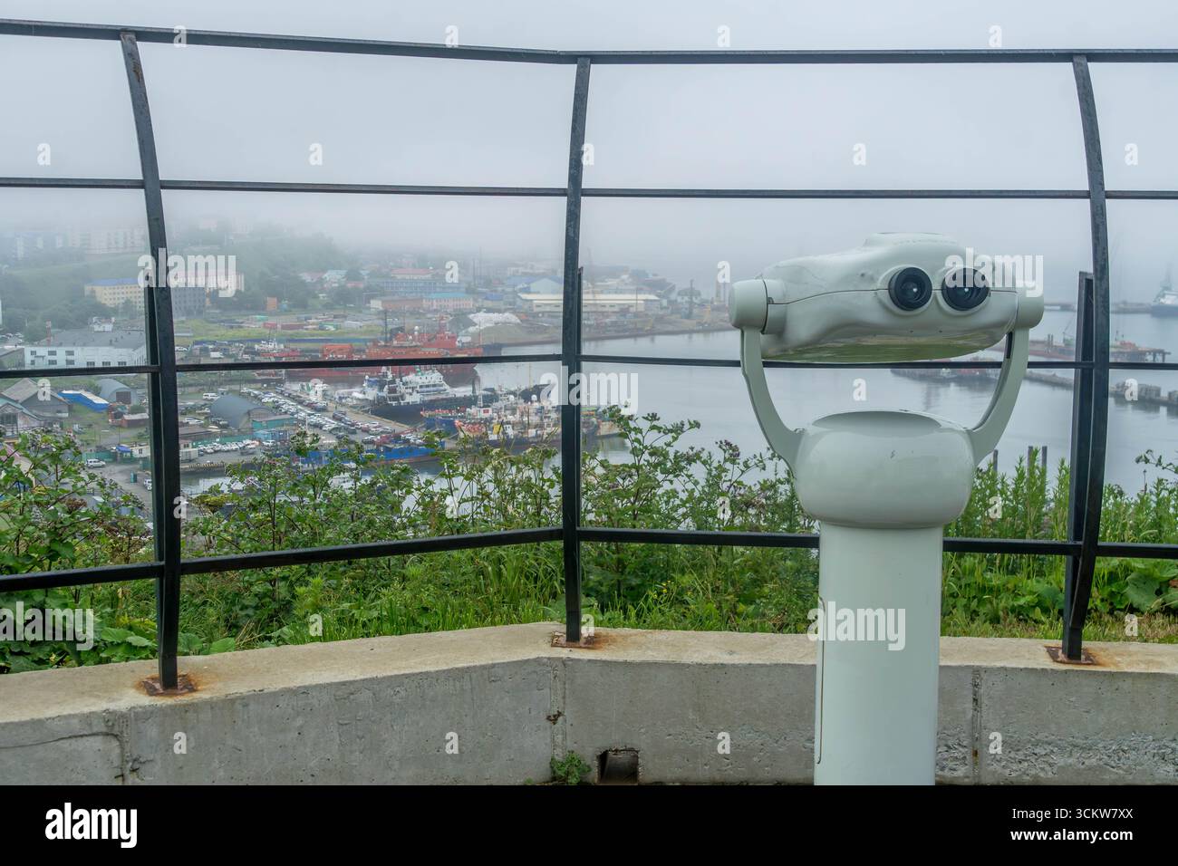 Visionneuse jumelle à pièces sur une colline surplombant le port brumeux et la ville de Korsakov, île de Sakhaline, Russie, avec des navires et des bâtiments industriels Banque D'Images