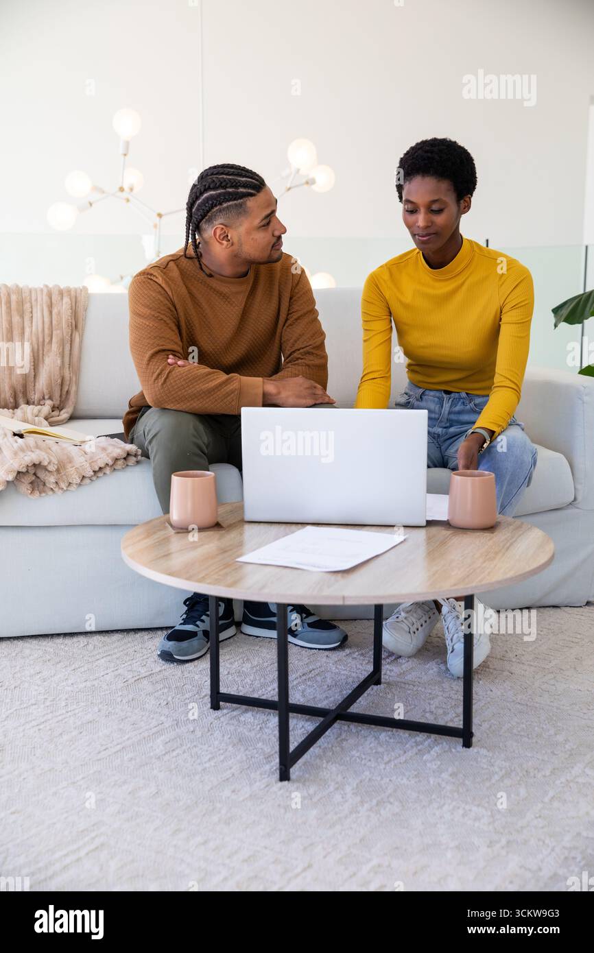 Couple diversifié collaborant sur le projet sur le canapé dans le salon avec le document d'ordinateur portable argenté et les tasses Banque D'Images Couple diversifié collaborant sur le projet sur le canapé dans le salon avec le document d'ordinateur portable argenté et les tasses Banque D'Images