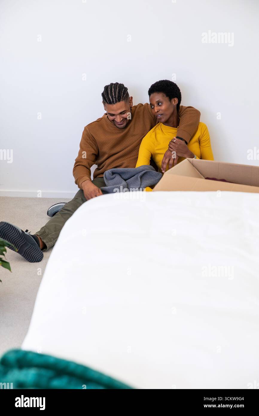 Couple diversifié assis sur la moquette dans la chambre déballant la boîte en carton avec couverture grise Banque D'Images