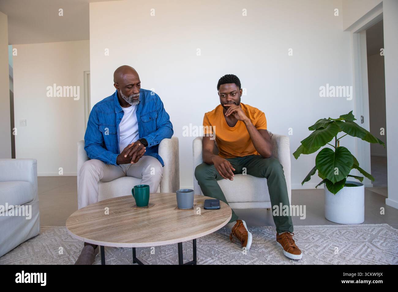 Hommes afro-américains sur des fauteuils à côté de la table à la maison sirotant des tasses près du smartphone Banque D'Images Hommes afro-américains sur des fauteuils à côté de la table à la maison sirotant des tasses près du smartphone Banque D'Images