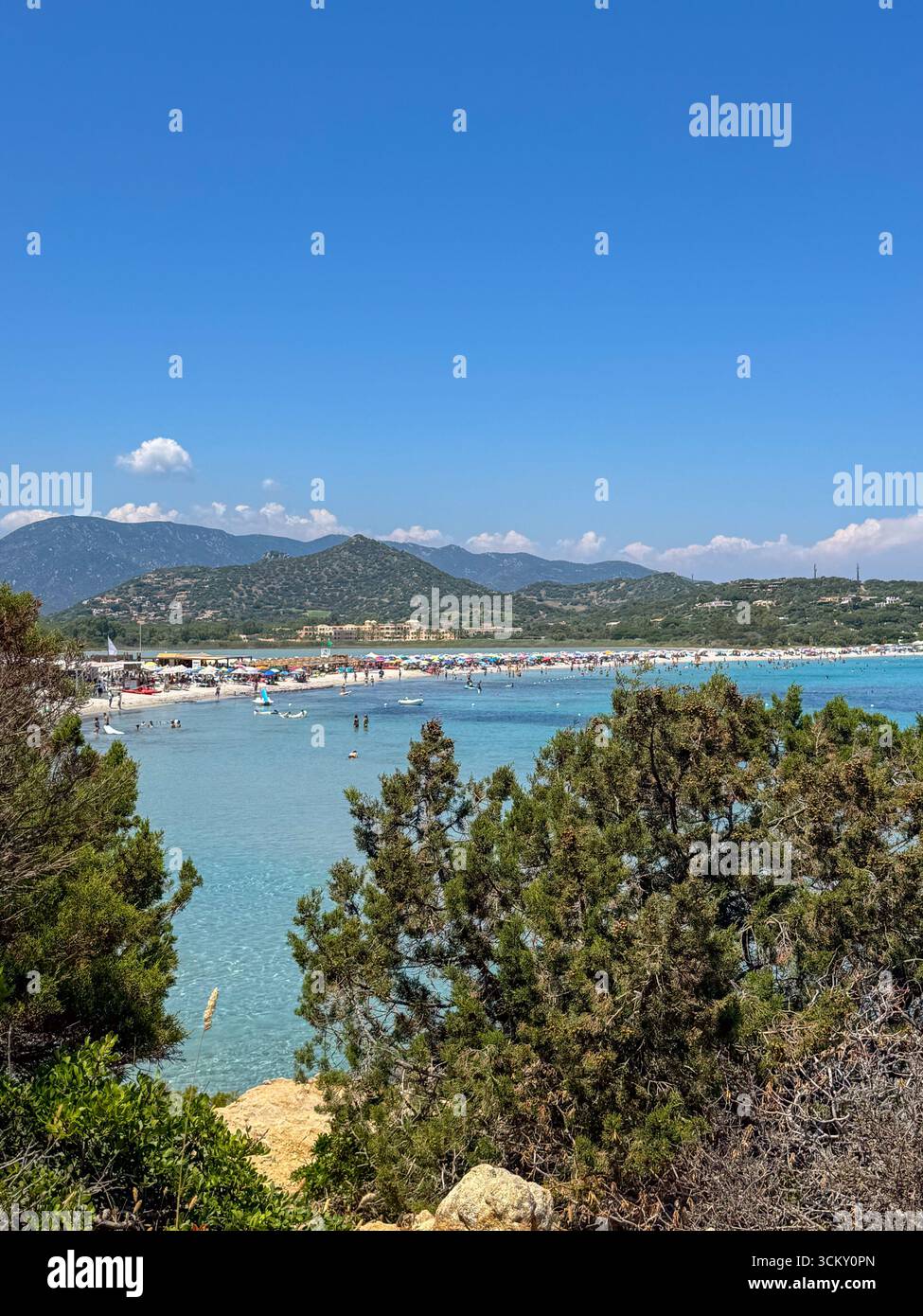 Journée d'été ensoleillée sur une plage populaire de Sardaigne avec des eaux turquoises, des bains de soleil et des montagnes vertes lointaines. Banque D'Images
