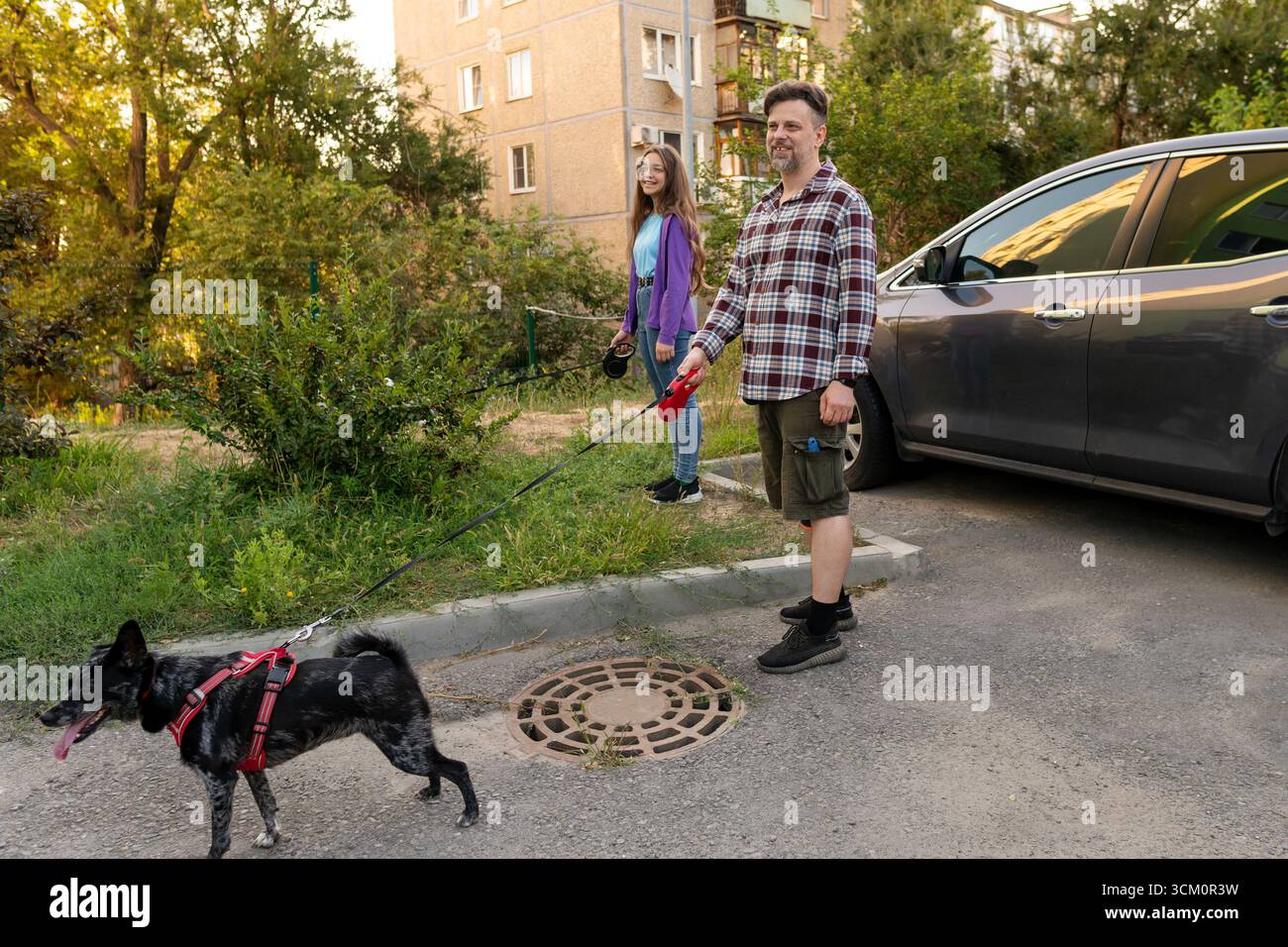 L'homme et sa fille sourient tout en promenant leur chien noir dans un harnais rouge près d'une voiture garée, tenant des laisses et profitant de la compagnie à l'extérieur dans un Banque D'Images