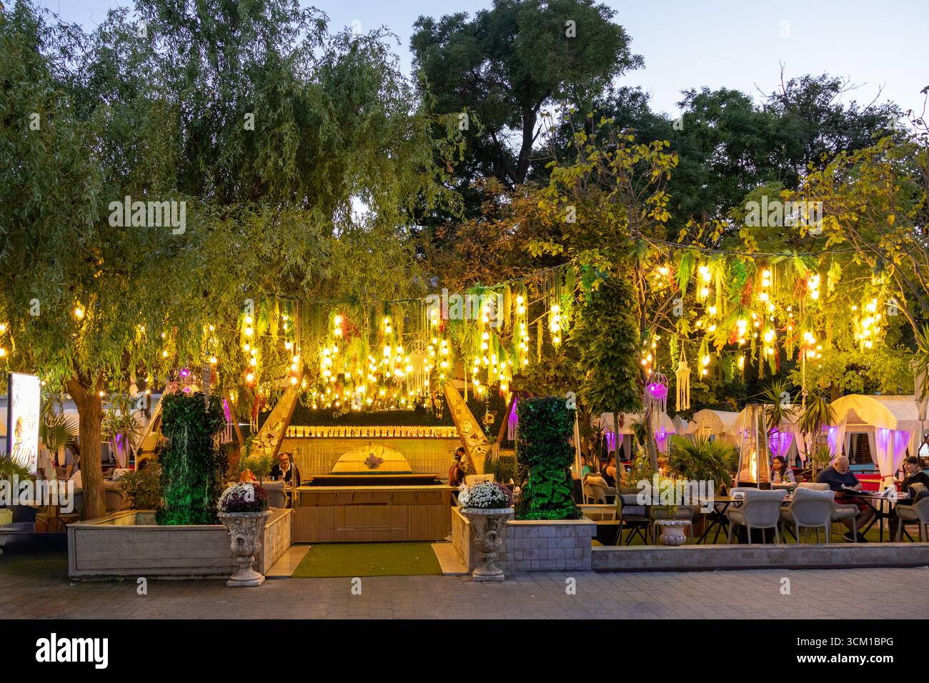 Vue en soirée sur une terrasse extérieure animée agrémentée de luminaires incandescents, d'arbres et d'un coin salon Banque D'Images
