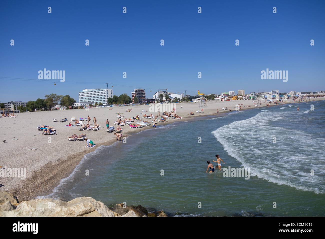 Scène de plage d'été animée avec des touristes nageant dans la mer, se relaxant sur le sable, et des hôtels en arrière-plan Banque D'Images