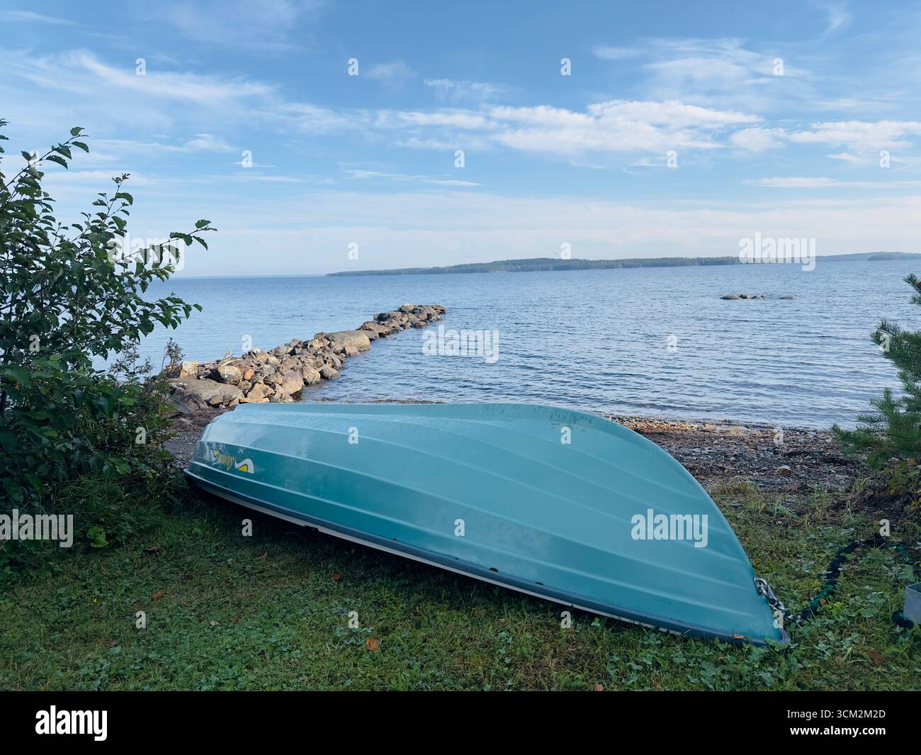 Un bateau sarcelle renversé repose sur la côte herbeuse près d'une jetée rocheuse et d'une eau calme. Banque D'Images