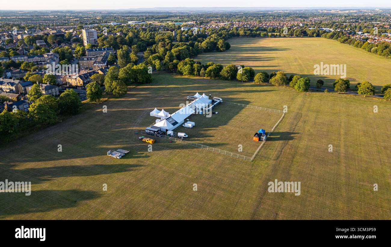 Une vue aérienne d'une grande structure de tente blanche avec des tentes plus petites, des véhicules et une zone clôturée sur un champ herbeux, avec une ville et des arbres dans le backgro Banque D'Images