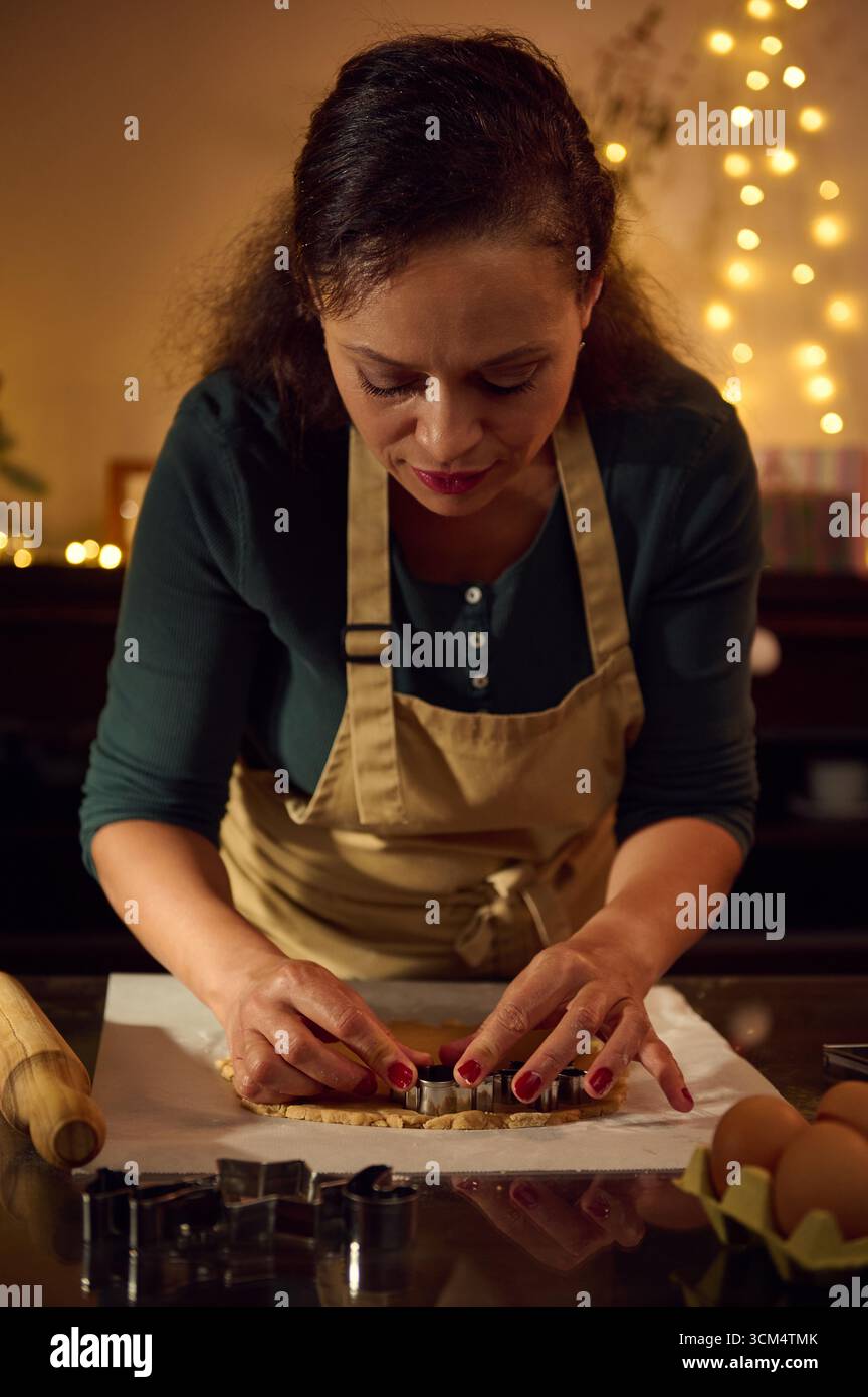 Une femme portant un tablier de chef prépare méticuleusement les biscuits des fêtes à l'aide d'emporte-pièce tout en étant entourée de lumières de Noël chaleureuses et festives, créant un c Banque D'Images