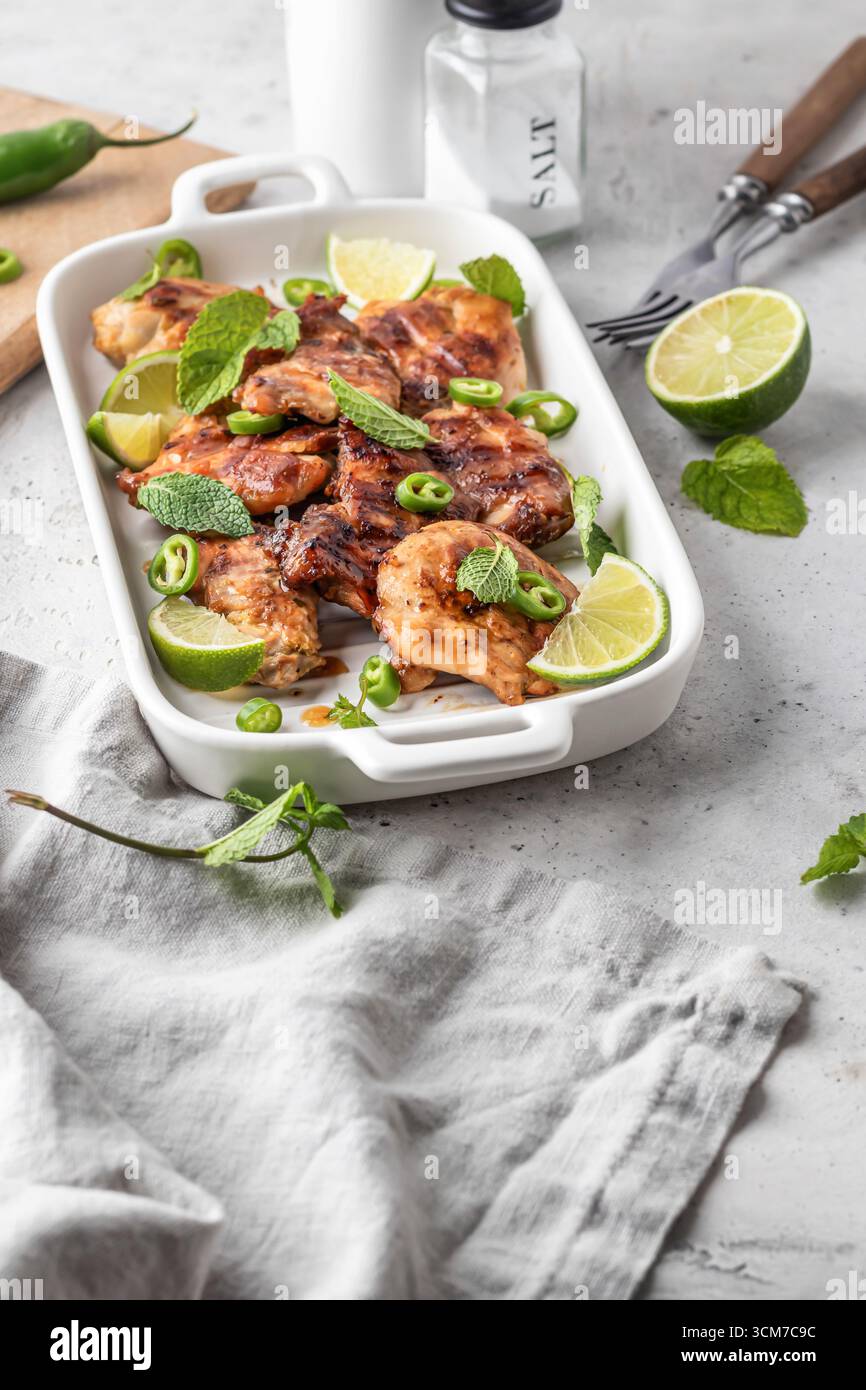 Cuisses de poulet grillées épicées rôties au piment et au citron vert sur plat blanc, fond de cuisine gris clair. Vertical Banque D'Images