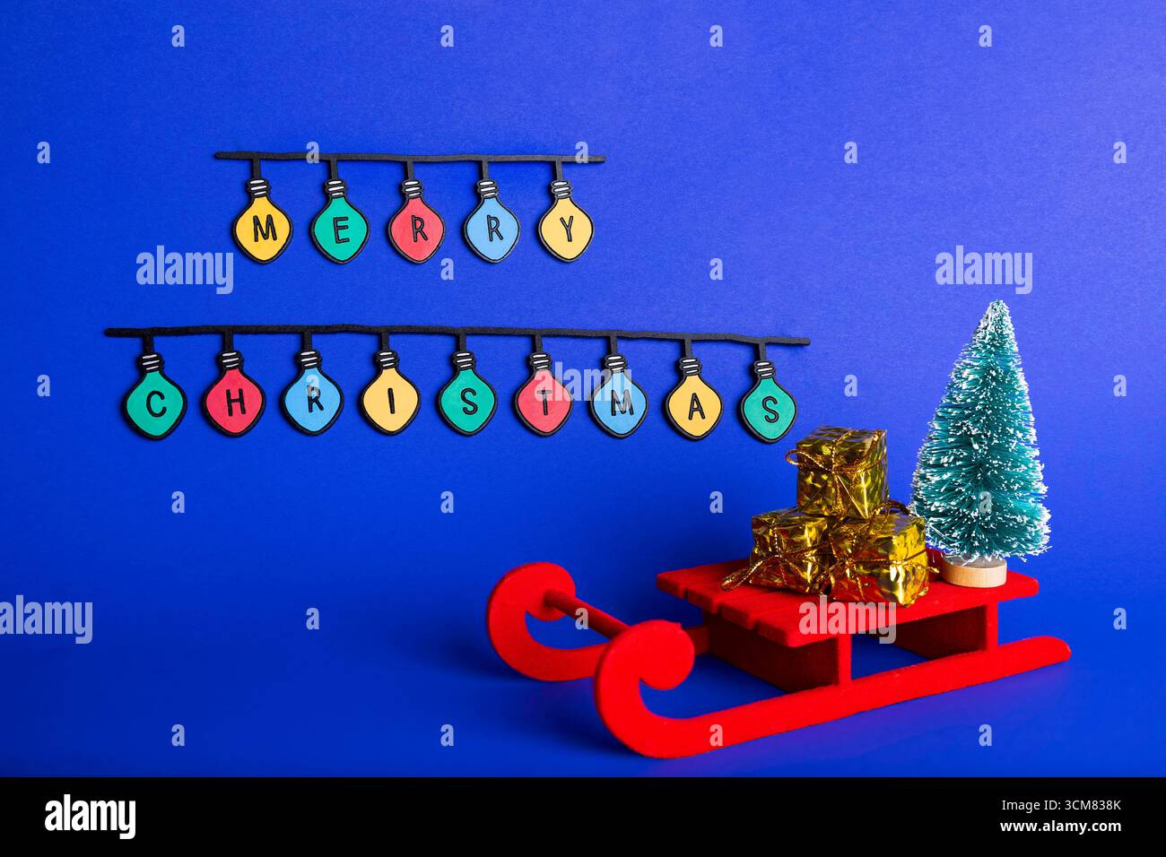 Composition créative de Noël avec un traîneau rouge avec des boîtes cadeaux dorées, un sapin de Noël vert miniature et des lumières colorées orthographiant « M » Banque D'Images