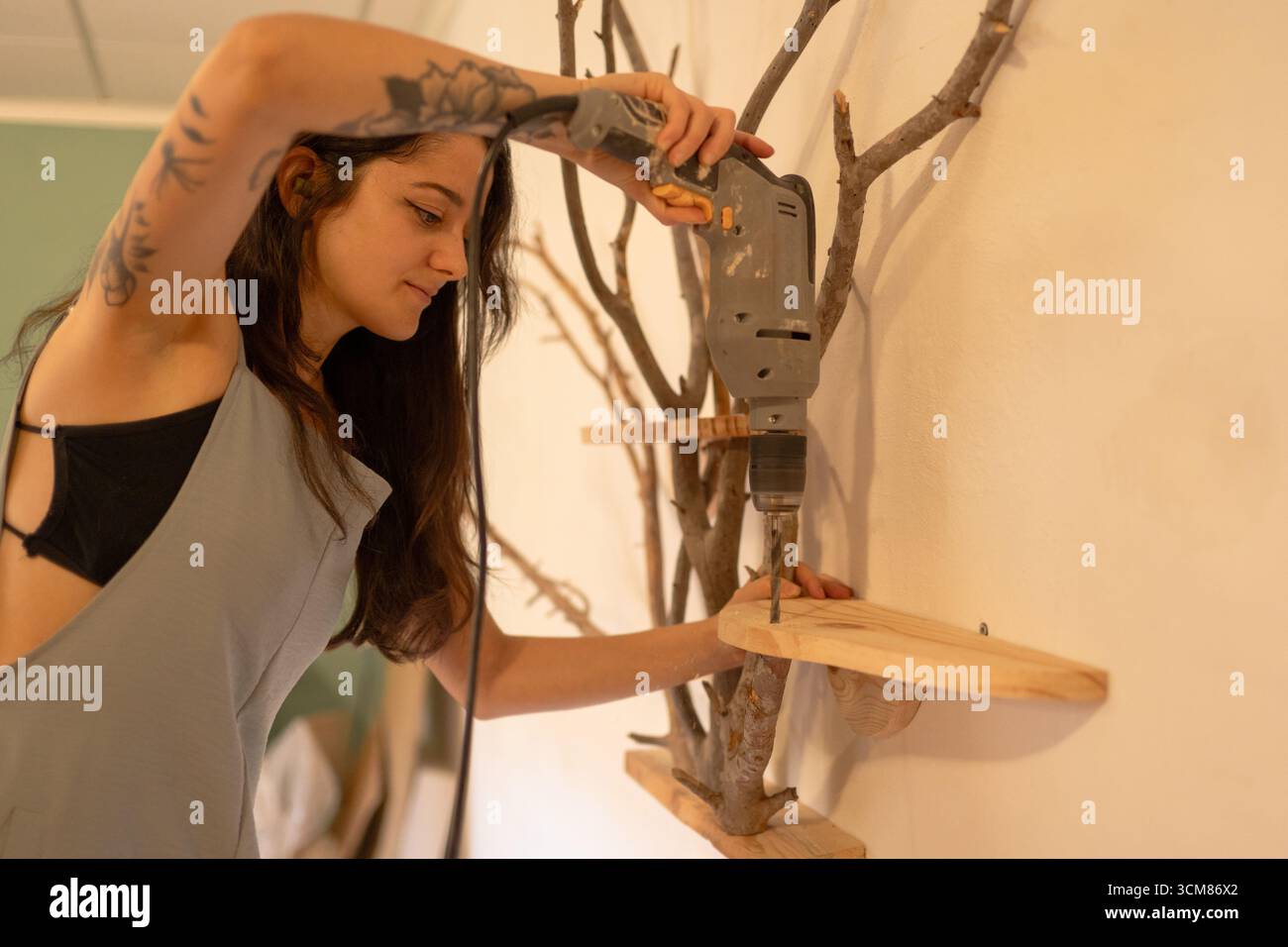 Femme utilisant la perceuse installant l'étagère en bois sur le décor rustique de mur de branche Banque D'Images