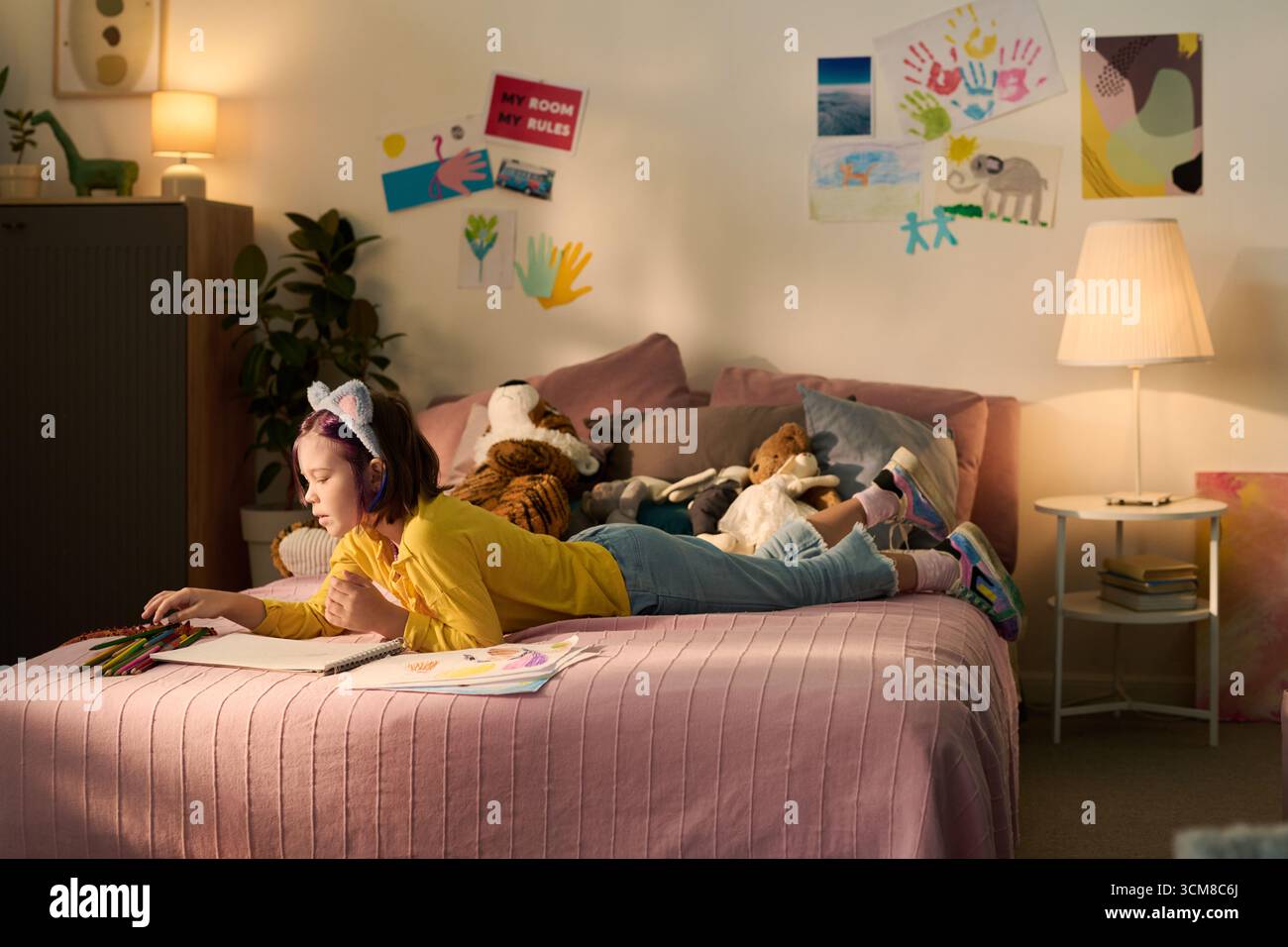 Fille caucasienne allongée sur le lit dessinant avec des crayons de couleur, entourée d'animaux en peluche et d'œuvres d'art pour enfants sur le mur, se concentrant sur l'activité créative dans le cadre de la chambre à coucher Banque D'Images