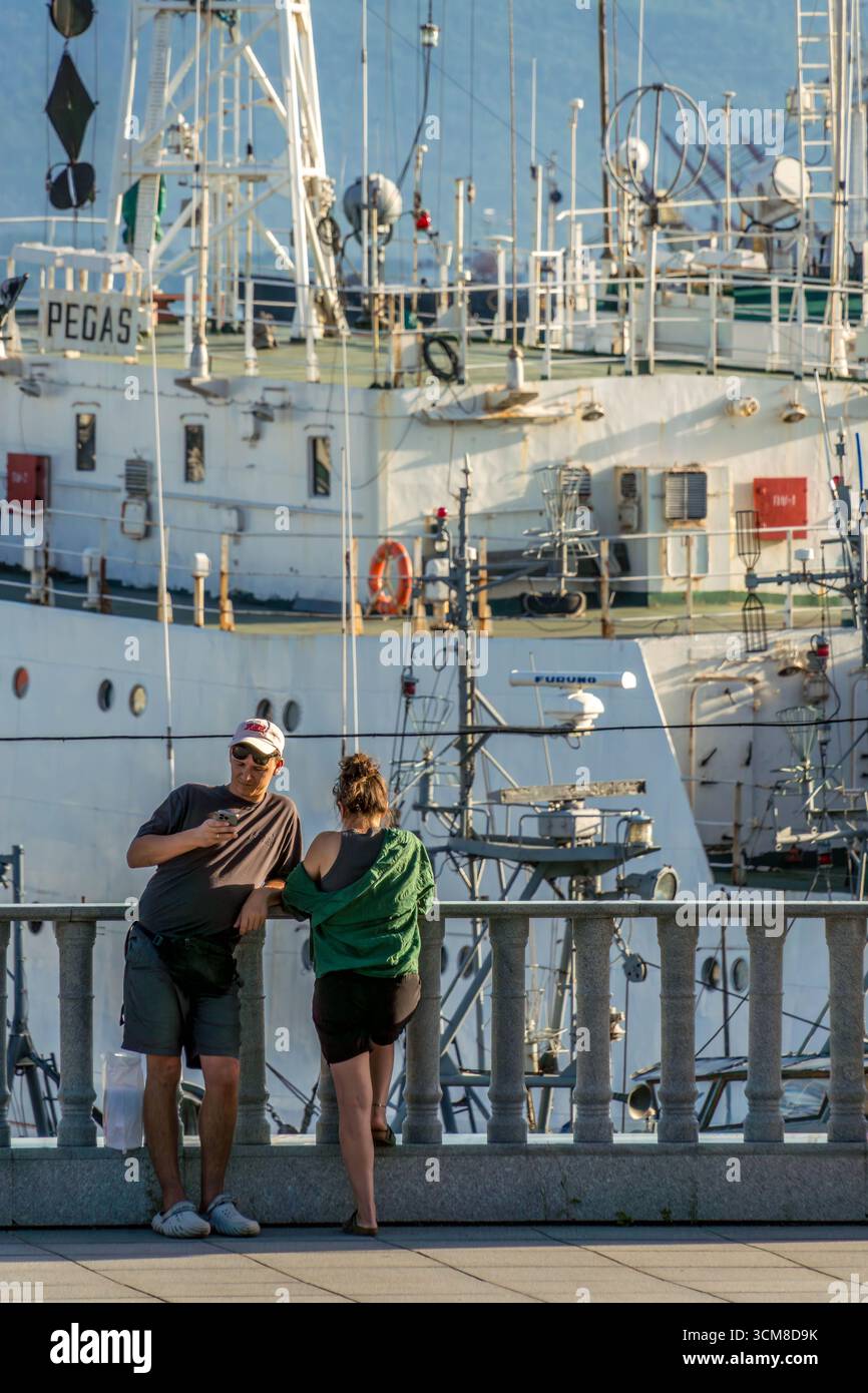 Deux personnes s'appuient sur une balustrade de pierre à Vladivostok, en Russie, avec le navire de recherche Pegas amarré. Banque D'Images