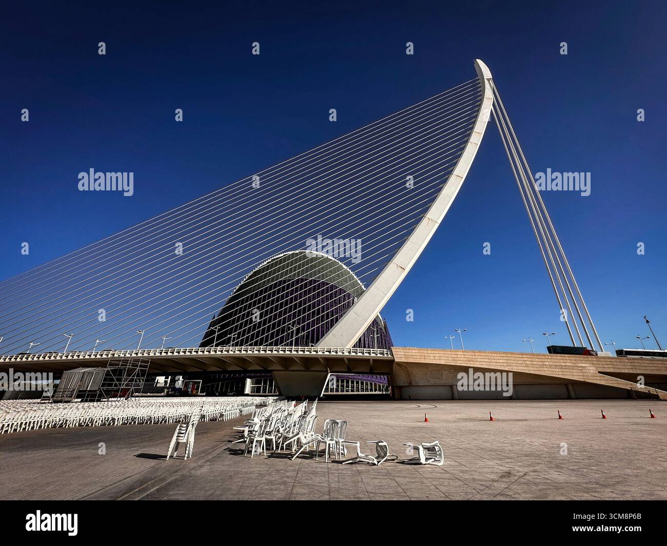 La photo montre l'impressionnant pont à haubans de Santiago Calatrava dans le cadre de la Ciudad de las Artes y las Ciencias à Valence, en Espagne. Au premier plan se trouvent de nombreuses chaises blanches, certaines disposées, certaines renversées, indiquant un événement extérieur à venir ou récemment terminé. Le contraste entre l’ingénierie moderne, le décor urbain et les objets du quotidien tels que les chaises confère à la scène une atmosphère particulière entre architecture et vie urbaine. Banque D'Images