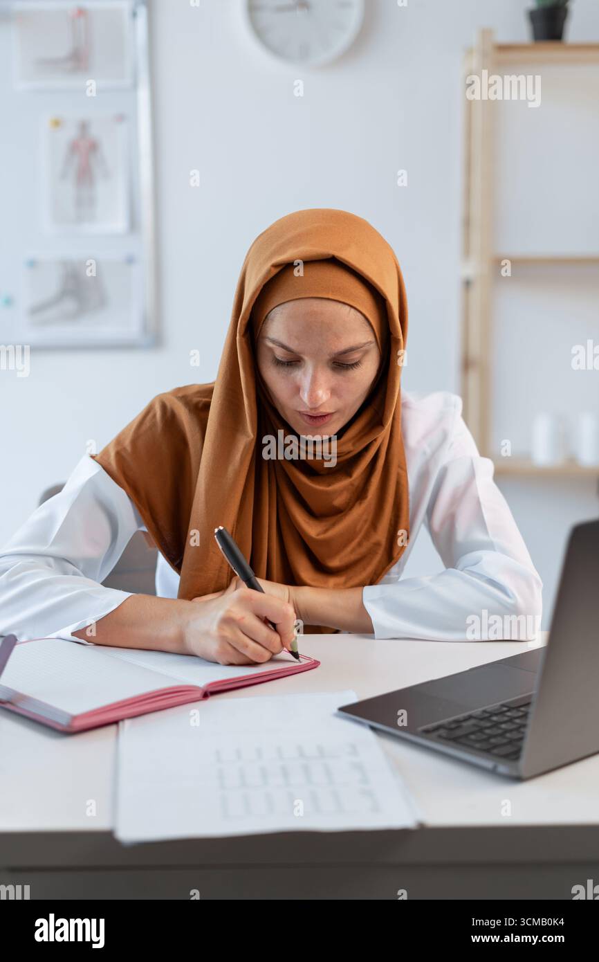 Médecin musulman féminin avec stéthoscope travaillant avec l'écriture de notes du patient et le diagnostic avec la concentration en clinique, en utilisant l'ordinateur portable dans l'hospi Banque D'Images