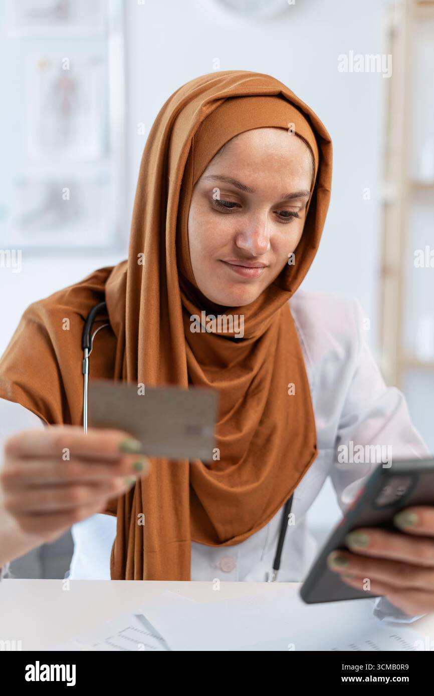 Médecin musulman en uniforme détenant une carte de crédit et utilisant un smartphone faisant des achats en ligne à l'hôpital. copier l'espace Banque D'Images
