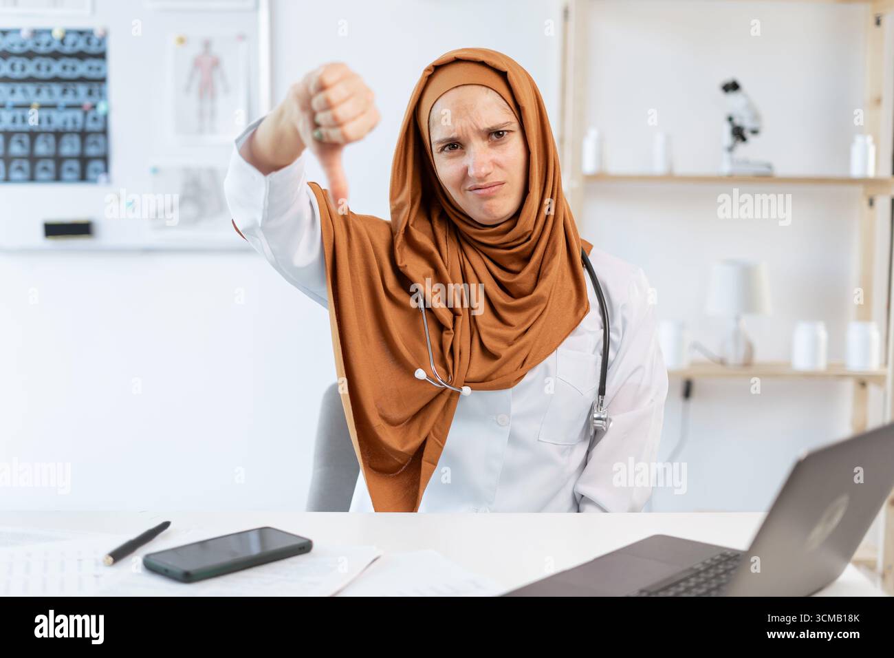 Triste femme musulmane médecin avec stéthoscope montrer le pouce vers le bas assis au bureau dans le bureau de l'hôpital. concept de médecine. rejet et émotions négatives. co Banque D'Images