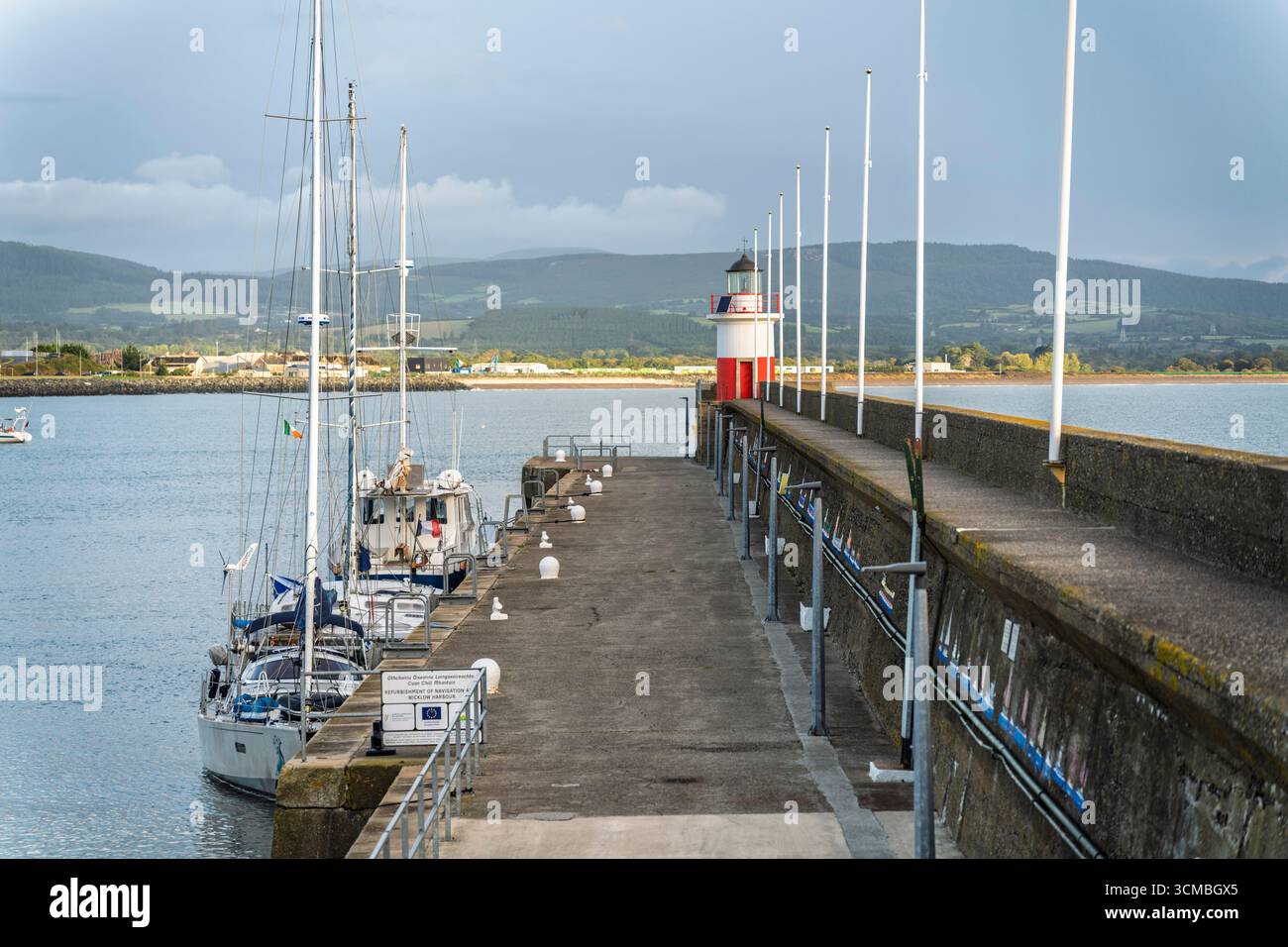 Jetée à Wicklow Harbour avec voilier amarré, phare rouge et blanc, et réflexions matinales sur un chemin en béton. Banque D'Images