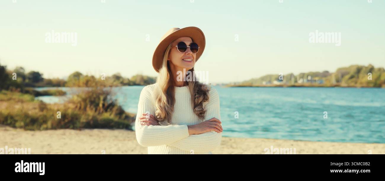 Femme mature élégante sur la plage côtière, modèle féminin d'âge moyen à la mode posant à l'extérieur, portant un pull, un chapeau et des lunettes, debout avec le bras croisé Banque D'Images