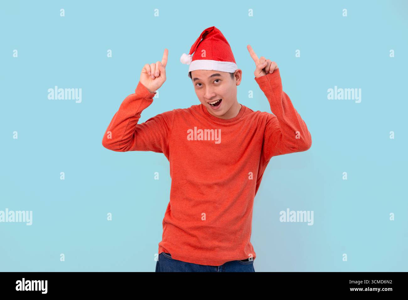 Un jeune homme joyeux portant un pull rouge et chapeau de Père Noël pointe vers le haut dans le concept de noël fond bleu Banque D'Images