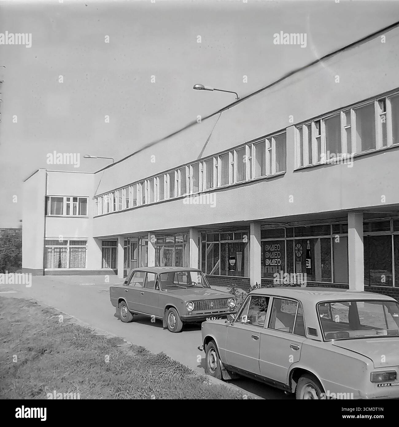 Une scène urbaine typique des dernières années de l'URSS est capturée dans cette photographie d'archives en noir et blanc de 1987 à Sloviansk, en RSS d'Ukraine. Une berline soviétique emblématique, la VAZ-2106 'Zhiguli', symbole de modeste prospérité à cette époque, est garée devant le restaurant 'Vesna' (printemps) dans le quartier résidentiel d'Artyom. Cette image sert de document historique précieux du mode de vie soviétique, des transports et de l'environnement urbain pendant la période charnière de la Perestroïka, mettant en valeur la vie quotidienne dans le contexte de l'architecture fonctionnaliste de l'époque Banque D'Images